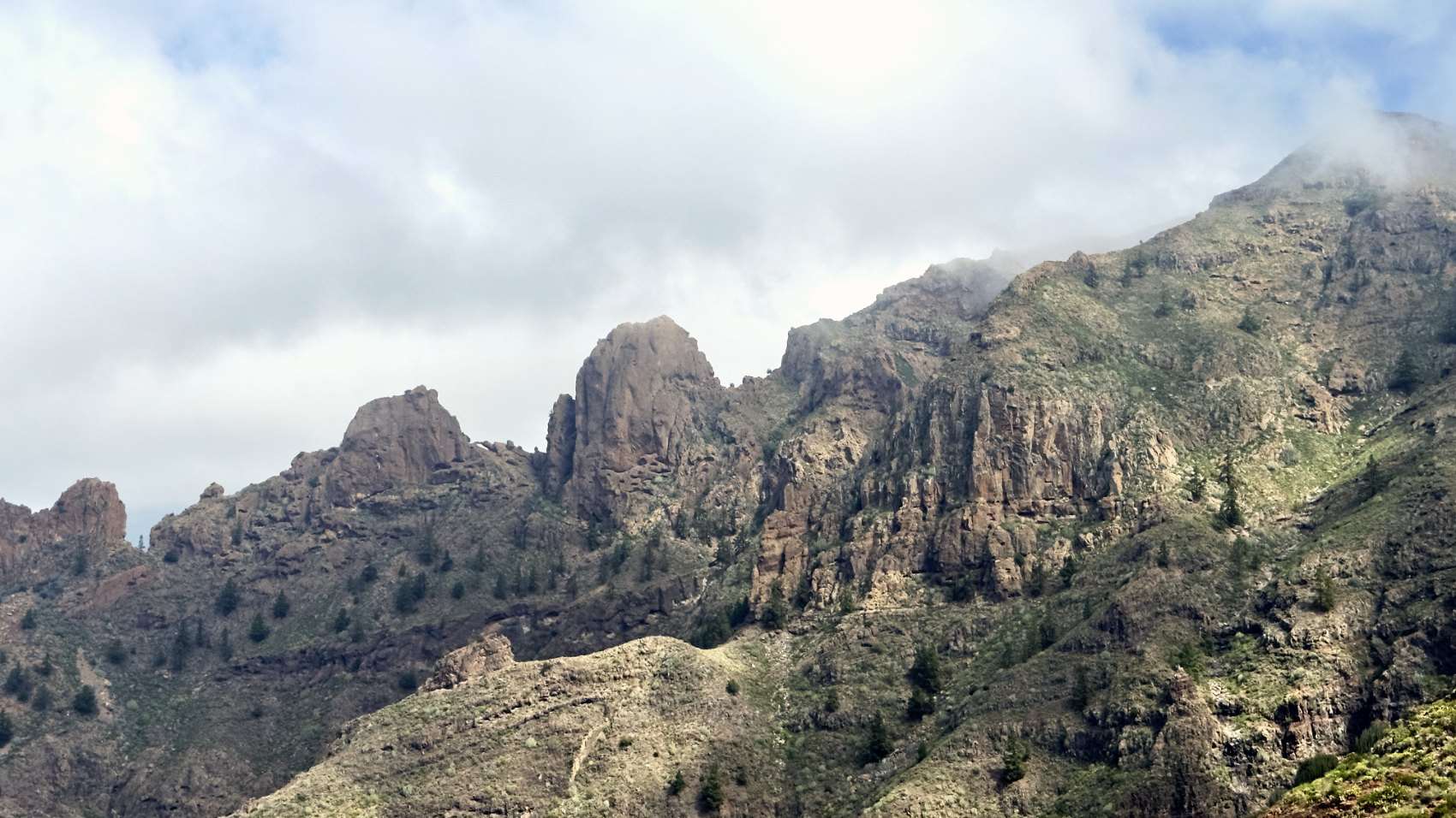 Rocky ridges and pinnacles above pine slopes at Ifonche, with cloud breaking over the crest.