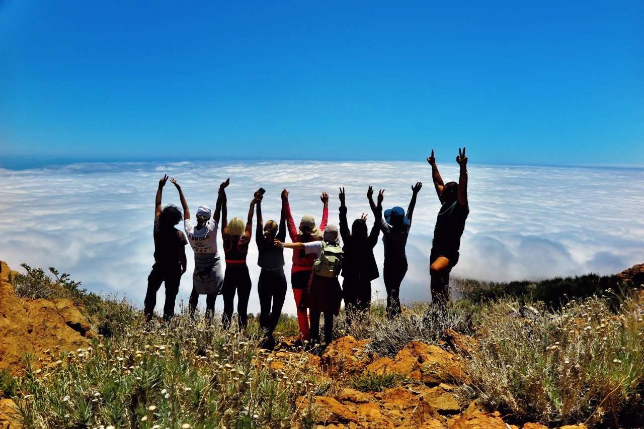 A group of walkers on a ridge with their arms raised, above an unbroken sea of cloud.