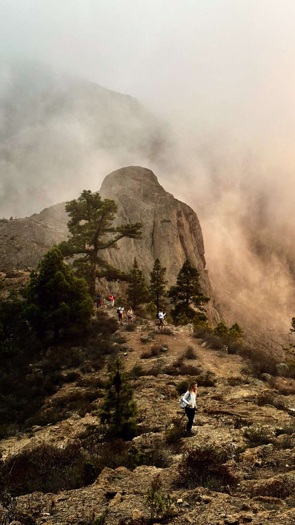 Walkers strung along a rocky ridge below a rounded rock face, with mist lit warm behind it.