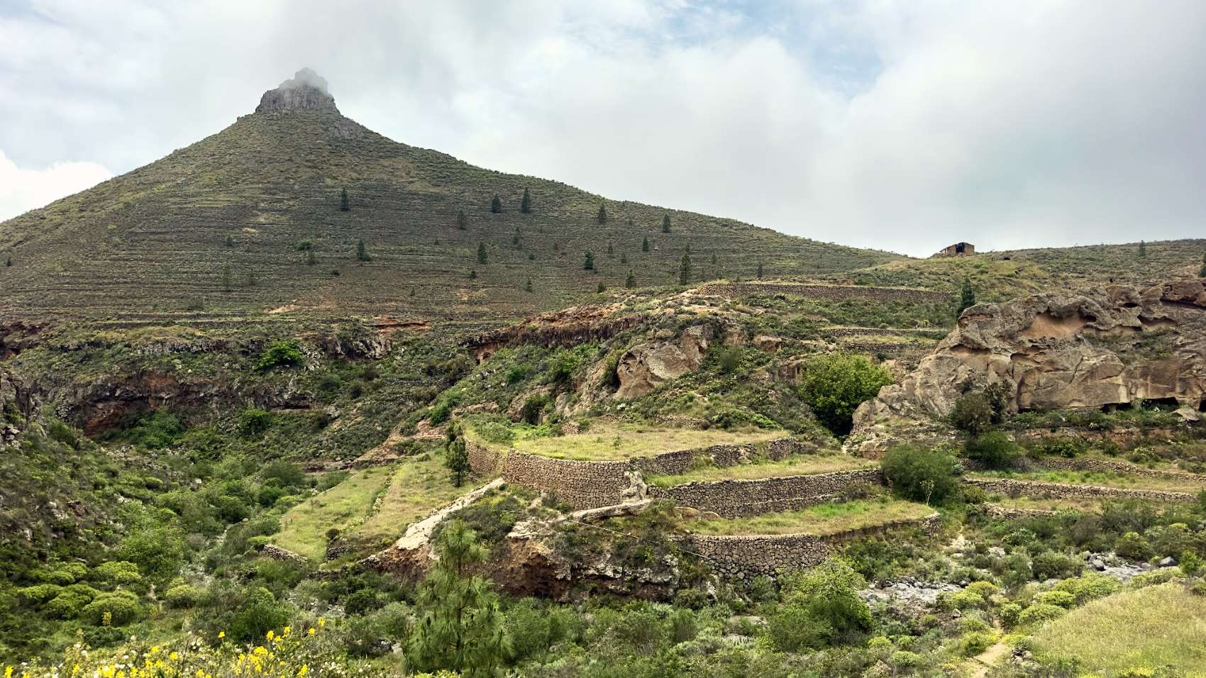 Dry-stone agricultural terraces below a conical peak at Ifonche, under a clouded sky.