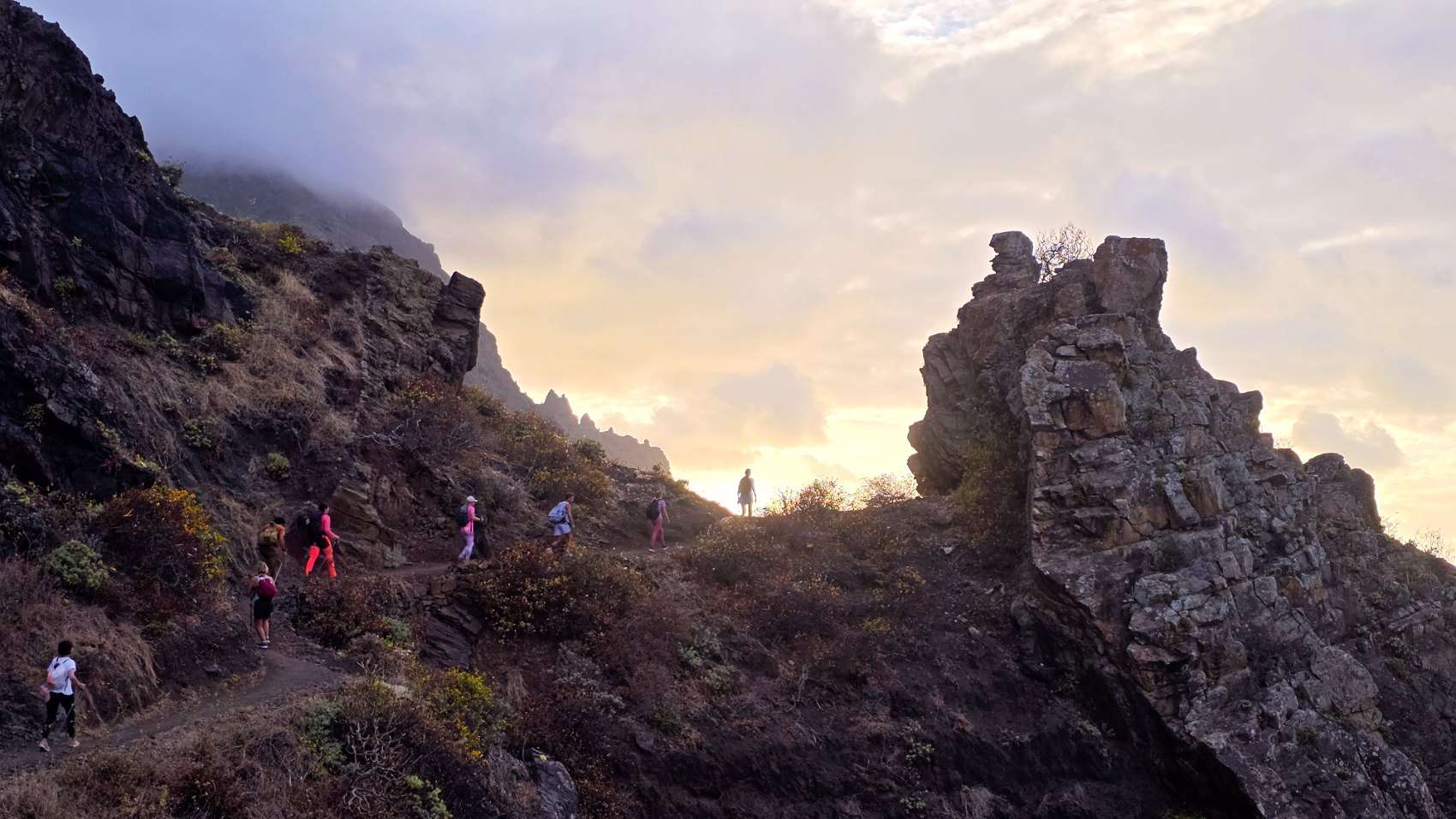Walkers on a ridge path in Anaga at sunset, a jagged rock tower standing to one side.
